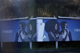 Two identical advertisement billboards display a man and a woman holding smartphones with broad smiles on their faces. Between them is an image of a smartphone with a pair of earbuds. The background is a dark blue, and the ads are placed outdoors with greenery visible behind.