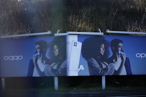 Two identical advertisement billboards display a man and a woman holding smartphones with broad smiles on their faces. Between them is an image of a smartphone with a pair of earbuds. The background is a dark blue, and the ads are placed outdoors with greenery visible behind.