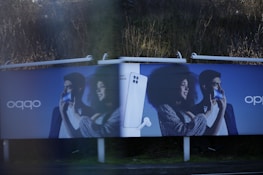 Two identical advertisement billboards display a man and a woman holding smartphones with broad smiles on their faces. Between them is an image of a smartphone with a pair of earbuds. The background is a dark blue, and the ads are placed outdoors with greenery visible behind.