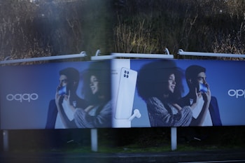 Two identical advertisement billboards display a man and a woman holding smartphones with broad smiles on their faces. Between them is an image of a smartphone with a pair of earbuds. The background is a dark blue, and the ads are placed outdoors with greenery visible behind.