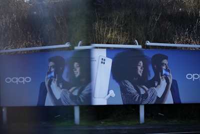 Two identical advertisement billboards display a man and a woman holding smartphones with broad smiles on their faces. Between them is an image of a smartphone with a pair of earbuds. The background is a dark blue, and the ads are placed outdoors with greenery visible behind.