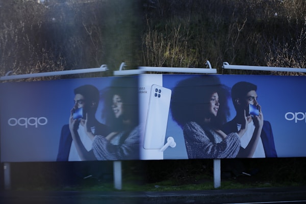 Two identical advertisement billboards display a man and a woman holding smartphones with broad smiles on their faces. Between them is an image of a smartphone with a pair of earbuds. The background is a dark blue, and the ads are placed outdoors with greenery visible behind.