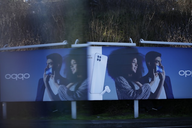 Two identical advertisement billboards display a man and a woman holding smartphones with broad smiles on their faces. Between them is an image of a smartphone with a pair of earbuds. The background is a dark blue, and the ads are placed outdoors with greenery visible behind.