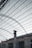 The image features a structure with an expansive metal framework, resembling an industrial or warehouse setting. The roof is open, revealing a network of beams and wires. Below, there is an old, weathered building with rusted metal bars and a tall chimney protruding through the roof.