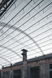 The image features a structure with an expansive metal framework, resembling an industrial or warehouse setting. The roof is open, revealing a network of beams and wires. Below, there is an old, weathered building with rusted metal bars and a tall chimney protruding through the roof.
