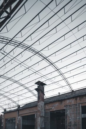 The image features a structure with an expansive metal framework, resembling an industrial or warehouse setting. The roof is open, revealing a network of beams and wires. Below, there is an old, weathered building with rusted metal bars and a tall chimney protruding through the roof.