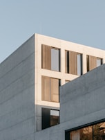 A modern concrete building with large windows and perforated metal panels. The structure has a minimalist design, featuring clean lines and a light facade under a clear blue sky.