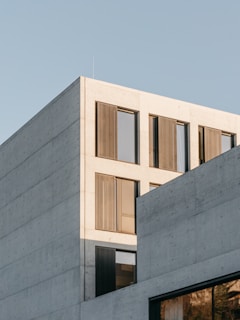 A modern concrete building with large windows and perforated metal panels. The structure has a minimalist design, featuring clean lines and a light facade under a clear blue sky.