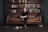 A young person sits on a brown leather couch in a library, surrounded by books. The bookshelf behind is filled with various books, and some books are scattered on the floor. The setting has a classic and cozy library feel.
