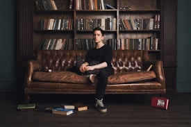 A young person sits on a brown leather couch in a library, surrounded by books. The bookshelf behind is filled with various books, and some books are scattered on the floor. The setting has a classic and cozy library feel.