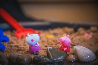 Two small cartoon pig figurines are placed on sandy soil, surrounded by various smooth stones. A bright red toy shovel and a blue toy element are partially visible in the background. The setting resembles a playful, miniature outdoor scene.