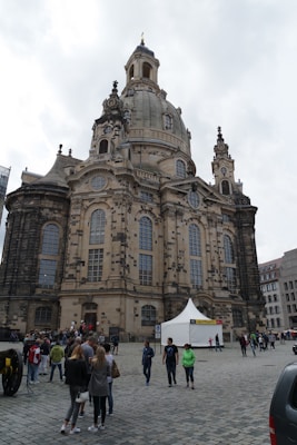 A large, historical baroque-style cathedral with intricate architectural details, including a prominent dome and multiple arched windows. People are walking in the cobblestone courtyard, and a white tent is set up nearby. The sky is overcast, adding contrast to the structure's stone facade.