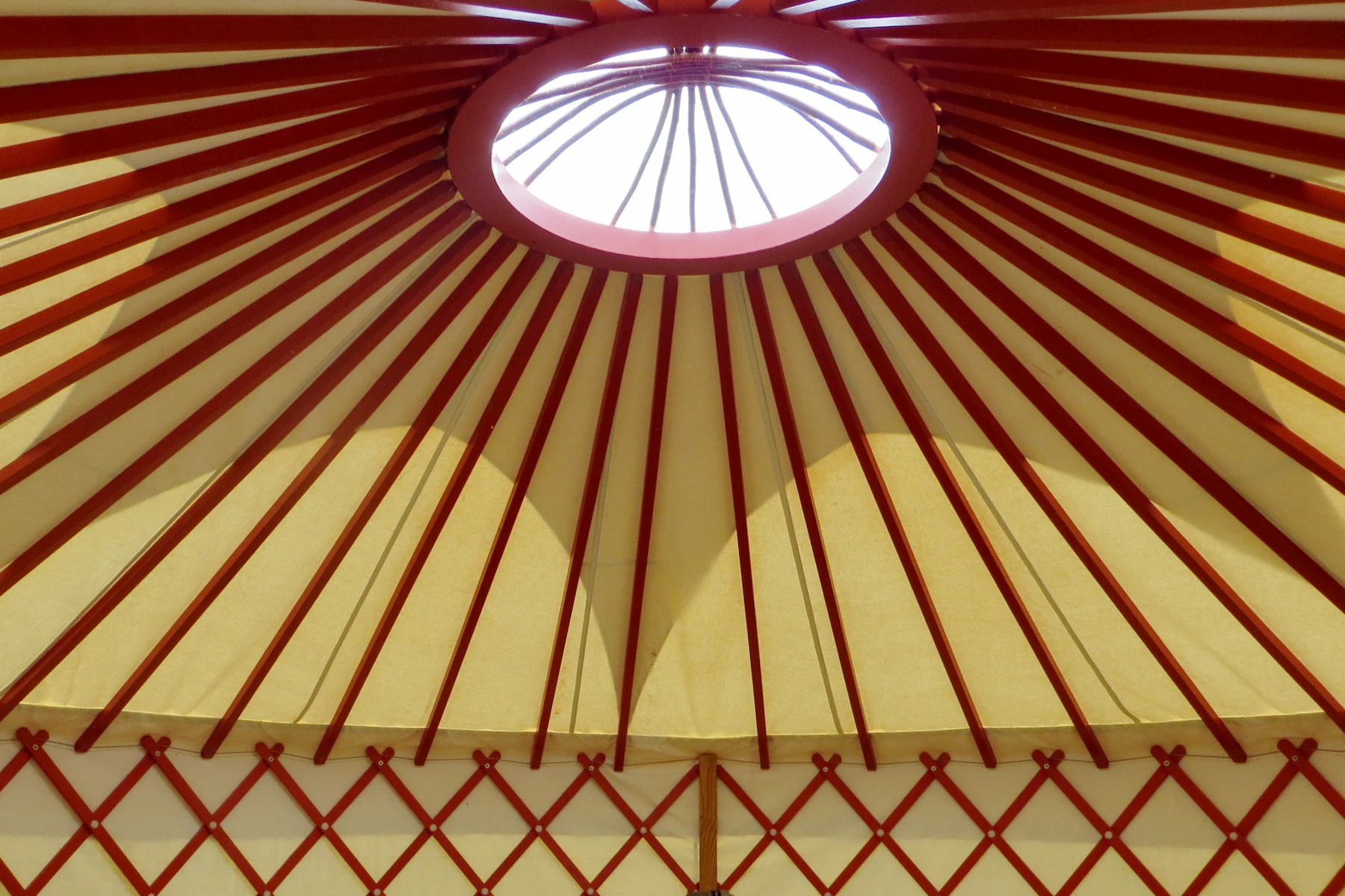 Looking up inside a yurt at red rafters fanning out from the roof ring to a red lattice wall, with cream fabric between them