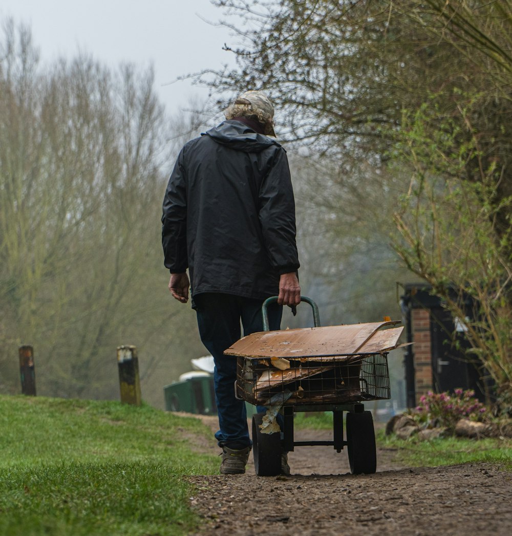 man in black jacket and brown hat standing in front of brown and black wheelbarrow