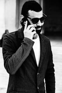 A man with a beard wearing sunglasses and a dark blazer is engaged in a phone call. He is holding a smartphone up to his ear. The background is blurred, suggesting he is outdoors or in a public place.