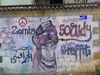 A graffiti mural on a wall featuring a stylized depiction of a zombie-like character with exaggerated features. The artwork includes bold, colorful text such as 'Zomby' and 'Graffiti' alongside Arabic script. There's a peace symbol and other artistic elements scattered throughout, with vibrant and dripping paint effects that add texture.