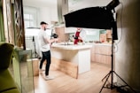 A kitchen setting is set up for a video or photo shoot. A man in a white shirt and dark jeans holds a camera, standing next to a well-lit kitchen island. Another person, wearing a maroon apron and gloves, appears to be preparing food. Studio lighting equipment surrounds the area, contributing to a bright, professional setup. The kitchen has wooden flooring and modern cabinetry.