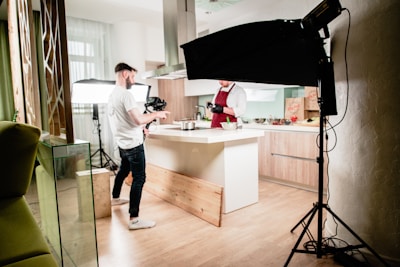 A kitchen setting is set up for a video or photo shoot. A man in a white shirt and dark jeans holds a camera, standing next to a well-lit kitchen island. Another person, wearing a maroon apron and gloves, appears to be preparing food. Studio lighting equipment surrounds the area, contributing to a bright, professional setup. The kitchen has wooden flooring and modern cabinetry.