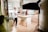 A kitchen setting is set up for a video or photo shoot. A man in a white shirt and dark jeans holds a camera, standing next to a well-lit kitchen island. Another person, wearing a maroon apron and gloves, appears to be preparing food. Studio lighting equipment surrounds the area, contributing to a bright, professional setup. The kitchen has wooden flooring and modern cabinetry.