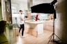 A kitchen setting is set up for a video or photo shoot. A man in a white shirt and dark jeans holds a camera, standing next to a well-lit kitchen island. Another person, wearing a maroon apron and gloves, appears to be preparing food. Studio lighting equipment surrounds the area, contributing to a bright, professional setup. The kitchen has wooden flooring and modern cabinetry.