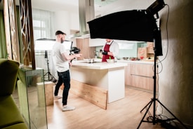 A kitchen setting is set up for a video or photo shoot. A man in a white shirt and dark jeans holds a camera, standing next to a well-lit kitchen island. Another person, wearing a maroon apron and gloves, appears to be preparing food. Studio lighting equipment surrounds the area, contributing to a bright, professional setup. The kitchen has wooden flooring and modern cabinetry.