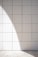 A wall of white tiles with a subtle geometric pattern from their arrangement. Sunlight casts a soft, diagonal shadow across the surface, creating a gentle contrast between light and shadow. A tiled floor at the base reflects similar muted tones.