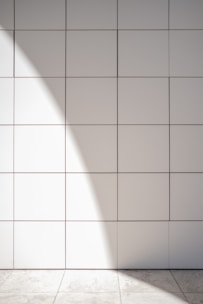 A wall of white tiles with a subtle geometric pattern from their arrangement. Sunlight casts a soft, diagonal shadow across the surface, creating a gentle contrast between light and shadow. A tiled floor at the base reflects similar muted tones.