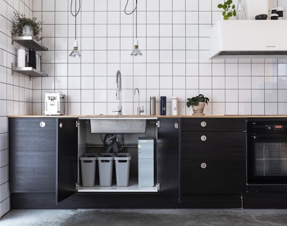 A modern kitchen interior featuring minimalist black cabinets and white grid-tiled walls. There are hanging pendant lights and various kitchen appliances on the counter, including a coffee machine. A sink with a gooseneck faucet is centrally positioned above open cabinets housing recycling bins. A potted plant adds a touch of greenery.