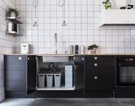 A modern kitchen interior featuring minimalist black cabinets and white grid-tiled walls. There are hanging pendant lights and various kitchen appliances on the counter, including a coffee machine. A sink with a gooseneck faucet is centrally positioned above open cabinets housing recycling bins. A potted plant adds a touch of greenery.