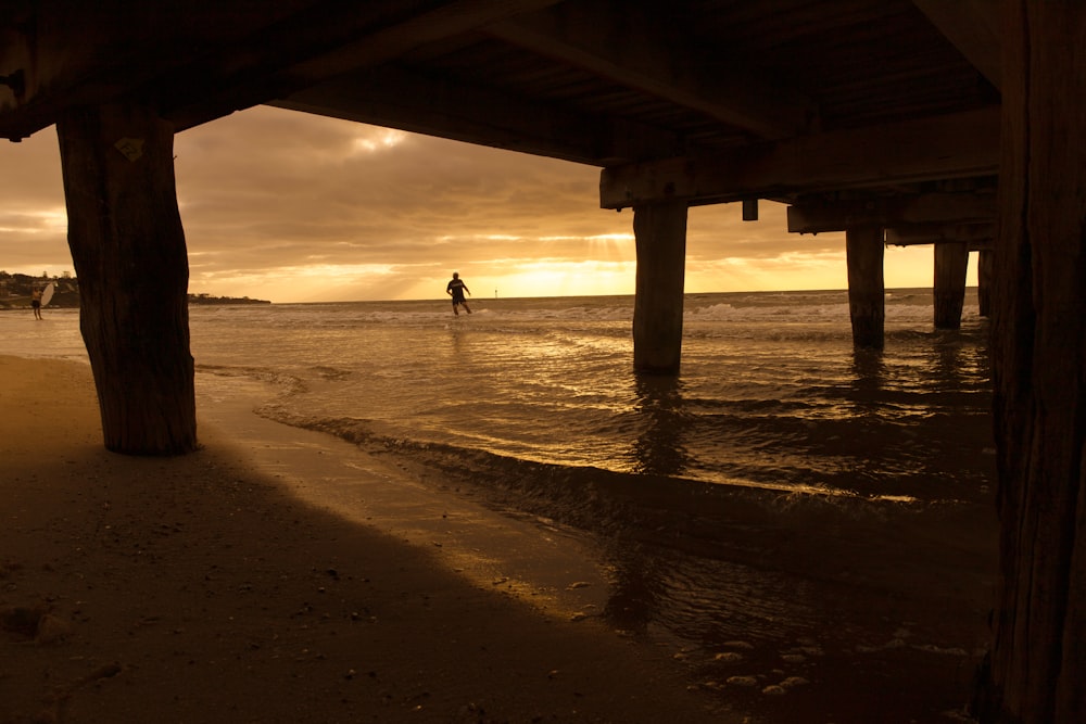 pessoa andando na praia sob a ponte de madeira durante o dia