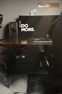 A workspace setup featuring a modern desktop with a large monitor displaying the text 'DO MORE.' A desktop speaker is positioned on the left side, and there is a keyboard with a mouse on the wooden desk. A pair of headphones is hanging under the desk, and a computer chair is partially visible on the right. A decorative item and a license plate with 'PZ8 UNIMOG' are mounted on the wall.