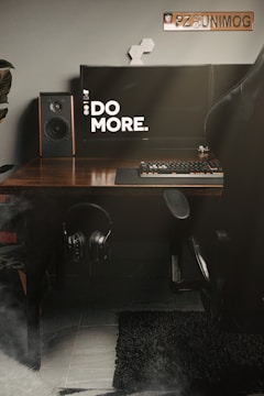 A workspace setup featuring a modern desktop with a large monitor displaying the text 'DO MORE.' A desktop speaker is positioned on the left side, and there is a keyboard with a mouse on the wooden desk. A pair of headphones is hanging under the desk, and a computer chair is partially visible on the right. A decorative item and a license plate with 'PZ8 UNIMOG' are mounted on the wall.