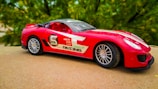 A red toy sports car with racing decals is placed on a flat surface. The car features a sleek design with black-tinted windows, silver rims, and various stickers such as 'Speed,' 'GTS line,' and 'Racing.' The background displays a blurred view of green foliage.
