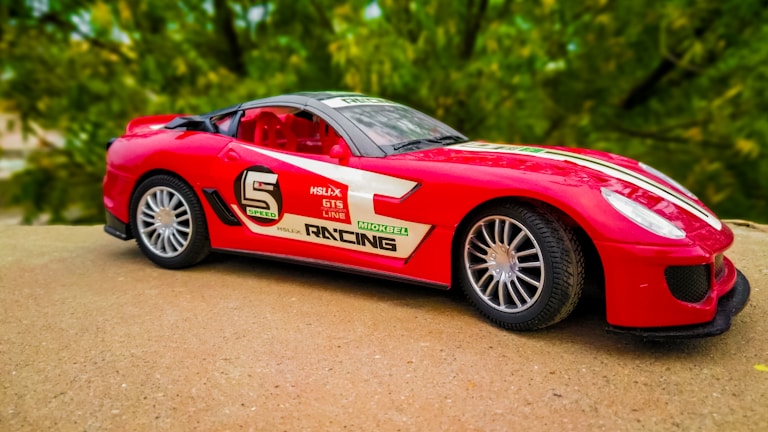 A red toy sports car with racing decals is placed on a flat surface. The car features a sleek design with black-tinted windows, silver rims, and various stickers such as 'Speed,' 'GTS line,' and 'Racing.' The background displays a blurred view of green foliage.