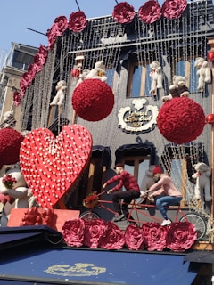 A decorated building facade features large red floral spheres, abundant red roses, and various teddy bears. Two mannequins are posed on a tandem bicycle in front of a giant red heart. Strings of small lights drape the building, creating a festive and romantic appearance.