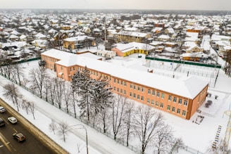 An aerial view of a snowy urban landscape featuring a large rectangular building with a pink facade and a white snow-covered roof. Surrounding the building are barren trees and a fenced sports area. The backdrop is a dense residential neighborhood with small houses, all capped with snow. A busy road with several cars runs along the front of the scene.
