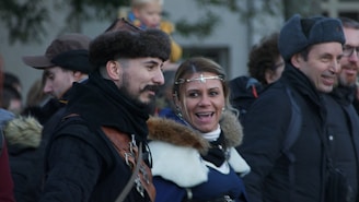A warm medieval festival scene with people in period costumes chatting around a wooden booth under colorful banners.