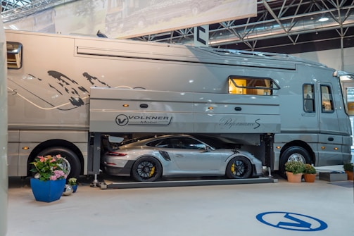 A large luxury motorhome with a built-in garage is showcased indoors. A high-end sports car is parked inside the garage compartment of the motorhome. There are potted plants arranged around the vehicle, and overhead lighting suggests an exhibition or show setting.