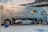 A large luxury motorhome with a built-in garage is showcased indoors. A high-end sports car is parked inside the garage compartment of the motorhome. There are potted plants arranged around the vehicle, and overhead lighting suggests an exhibition or show setting.