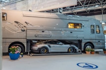 A large luxury motorhome with a built-in garage is showcased indoors. A high-end sports car is parked inside the garage compartment of the motorhome. There are potted plants arranged around the vehicle, and overhead lighting suggests an exhibition or show setting.