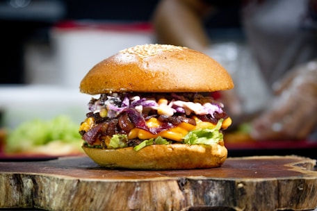 A gourmet burger sits on a wooden board, featuring a sesame seed bun. The burger is stacked with fresh green lettuce, crispy bacon, melted cheddar cheese, and a colorful mix of cabbage slaw topped with creamy sauce.