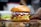 A gourmet burger sits on a wooden board, featuring a sesame seed bun. The burger is stacked with fresh green lettuce, crispy bacon, melted cheddar cheese, and a colorful mix of cabbage slaw topped with creamy sauce.