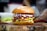 A gourmet burger sits on a wooden board, featuring a sesame seed bun. The burger is stacked with fresh green lettuce, crispy bacon, melted cheddar cheese, and a colorful mix of cabbage slaw topped with creamy sauce.