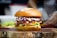 A gourmet burger sits on a wooden board, featuring a sesame seed bun. The burger is stacked with fresh green lettuce, crispy bacon, melted cheddar cheese, and a colorful mix of cabbage slaw topped with creamy sauce.