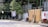 Clothing is hanging on a clothesline in an outdoor setting, with green foliage and shrubs along a white fence. There are various items scattered on the ground, including a wooden stool, metal barrels, and some construction materials. The background includes partly hidden structures with wooden and metallic elements.