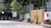 Clothing is hanging on a clothesline in an outdoor setting, with green foliage and shrubs along a white fence. There are various items scattered on the ground, including a wooden stool, metal barrels, and some construction materials. The background includes partly hidden structures with wooden and metallic elements.