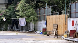 Clothing is hanging on a clothesline in an outdoor setting, with green foliage and shrubs along a white fence. There are various items scattered on the ground, including a wooden stool, metal barrels, and some construction materials. The background includes partly hidden structures with wooden and metallic elements.