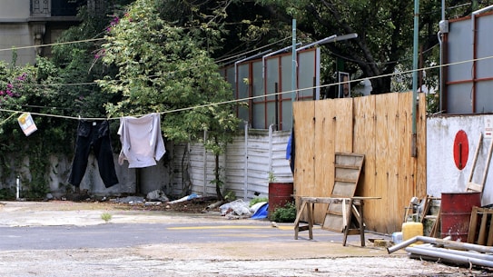 Clothing is hanging on a clothesline in an outdoor setting, with green foliage and shrubs along a white fence. There are various items scattered on the ground, including a wooden stool, metal barrels, and some construction materials. The background includes partly hidden structures with wooden and metallic elements.