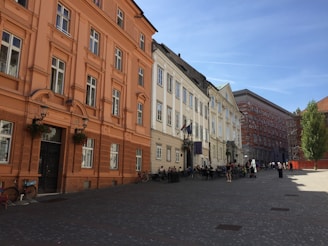 A city street lined with historic buildings featuring classical architectural styles. The buildings are painted in shades of orange, beige, and white. There are several people walking and sitting at outdoor cafes, and the scene is set under a bright blue sky. Bicycles are parked along the side of the street, adding to the casual, lively atmosphere.