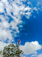 A telecommunications tower stands tall against a vibrant blue sky scattered with fluffy white clouds. Green foliage and overhead power lines frame the scene, adding a contrasting natural element to the industrial structure of the tower.